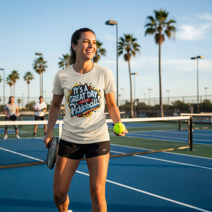 A woman playing pickleball on a court wearing a t-shirt that says It's A Great Day To Play Pickleball! with palm trees in the background.