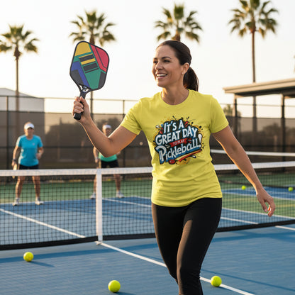 A woman playing pickleball on a court wearing a yellow t-shirt that says It's A Great Day To Play Pickleball! with palm trees in the background