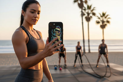 A woman taking a selfie with a LIVE FIT phone case on a beach with people exercising in the background.