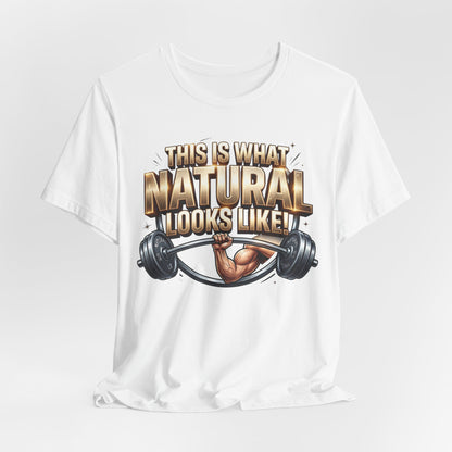 A white short-sleeve crewneck t-shirt with a graphic print on the front. The graphic features the text 'THIS IS WHAT NATURAL LOOKS LIKE!' in a bold, metallic gold and brown font, above an illustration of a muscular arm flexing a barbell.
