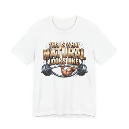 A white short-sleeve crewneck t-shirt featuring a graphic print on the front with the text 'THIS IS WHAT NATURAL LOOKS LIKE!' in a metallic gold font, above an illustration of a muscular arm lifting a barbell.
