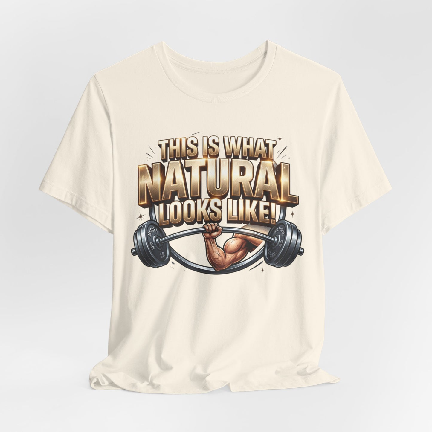 An off-white short-sleeve t-shirt with a graphic print on the front. The graphic features the text 'THIS IS WHAT NATURAL LOOKS LIKE!' in a bold, metallic gold and brown font, above an illustration of a muscular arm lifting a barbell.