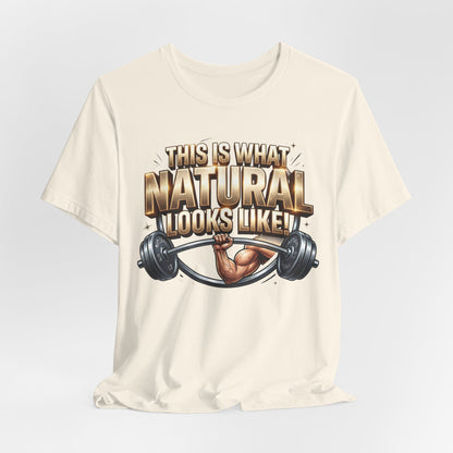 An off-white short-sleeve t-shirt with a graphic print on the front. The graphic features the text 'THIS IS WHAT NATURAL LOOKS LIKE!' in a bold, metallic gold and brown font, above an illustration of a muscular arm lifting a barbell.