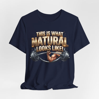 A navy blue short-sleeve t-shirt with a graphic print on the front. The graphic features the text 'THIS IS WHAT NATURAL LOOKS LIKE!' in a metallic gold and white font, above an illustration of a muscular arm lifting a barbell.