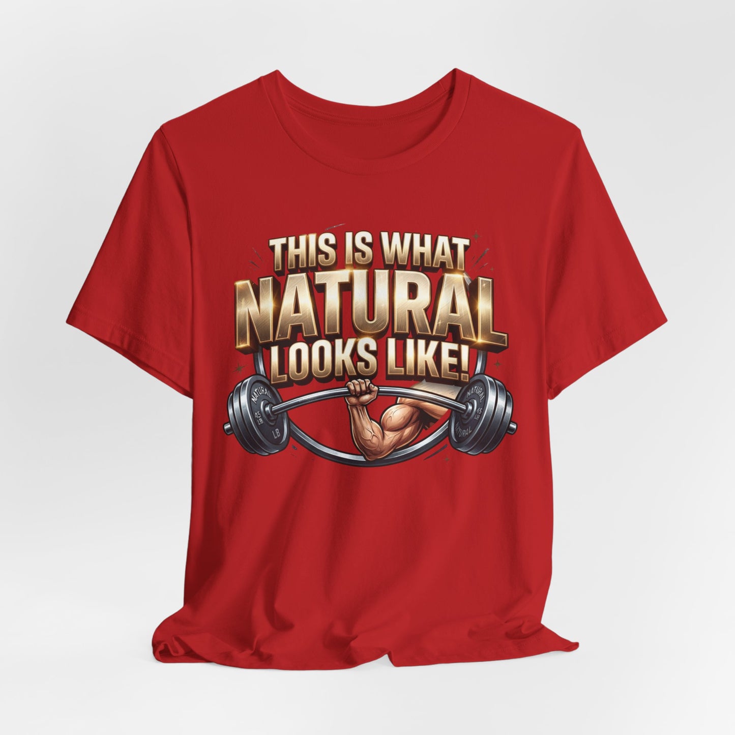 A red short-sleeve crewneck t-shirt with a graphic on the front that reads 'THIS IS WHAT NATURAL LOOKS LIKE!' in gold and white lettering, featuring an illustration of a muscular arm lifting a barbell.