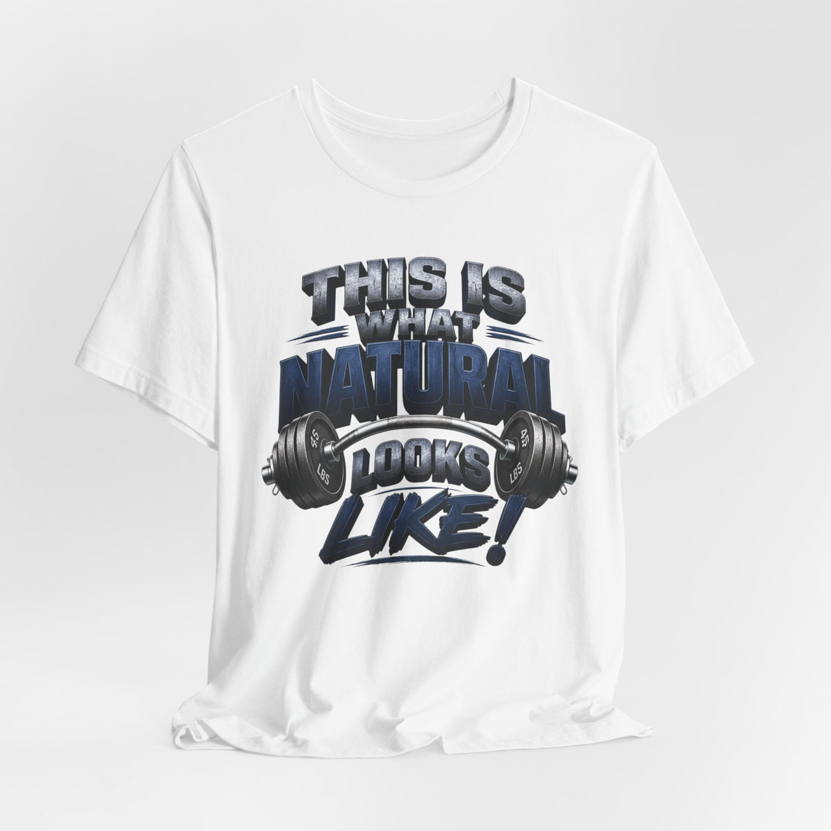 A white short-sleeve crewneck t-shirt with a graphic print on the front that reads 'THIS IS WHAT NATURAL LOOKS LIKE!' in a bold, stylized dark blue and gray font, accompanied by a barbell graphic. The t-shirt is displayed against a light gray studio background.