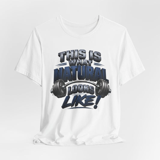 A white short-sleeve crewneck t-shirt with a graphic print on the front that reads 'THIS IS WHAT NATURAL LOOKS LIKE!' in a bold, stylized dark blue and gray font, accompanied by a barbell graphic. The t-shirt is displayed against a light gray studio background.
