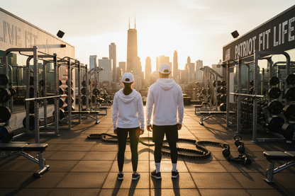 Two people in white hoodies and black leggings stand on a rooftop gym floor facing a city skyline at sunset. The scene includes weight machines, dumbbells, resistance bands, and large wall signs reading 'LIVE FIT' and 'PATRIOT FIT LIFE'.